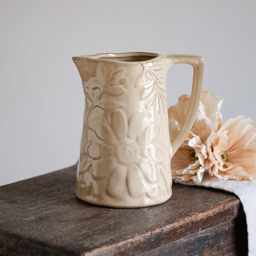 Beige ceramic pitcher with floral patterns on a wooden surface with flowers in the background