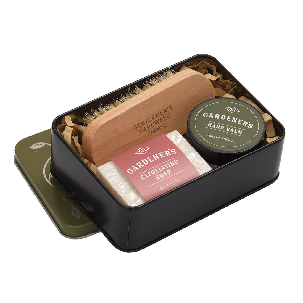 Set of gardening tools and products including a brush, soap, and balm in a black tin on a white background.