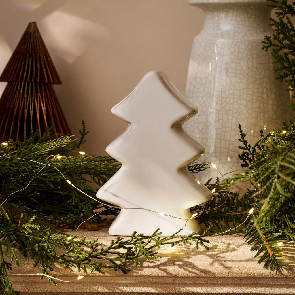 White ceramic Christmas tree ornament on a decorative surface with greenery and lights.