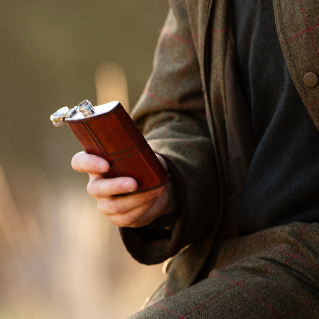 A person in a brown tweed jacket holds a small brown leather flask with a silver metal cap in their hand, set against a softly blurred outdoor background.