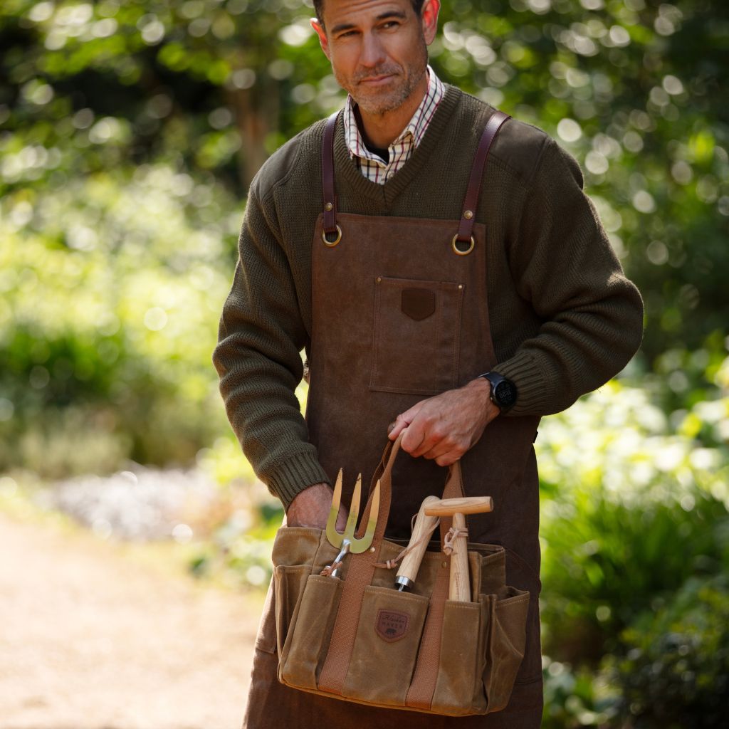 Man wearing a leather apron holding gardening tools in a garden bag