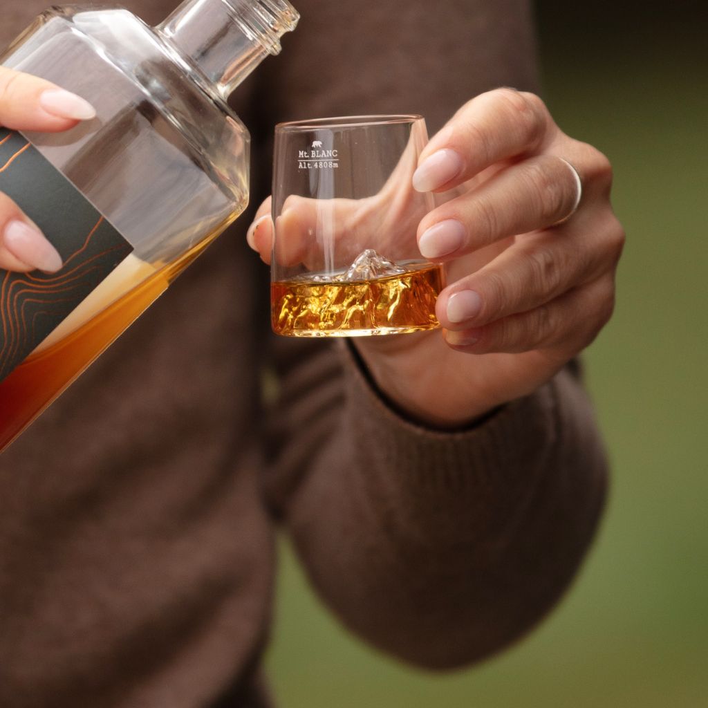 A person in a brown sweater pours amber liquid from a clear bottle into a topographic shot glass