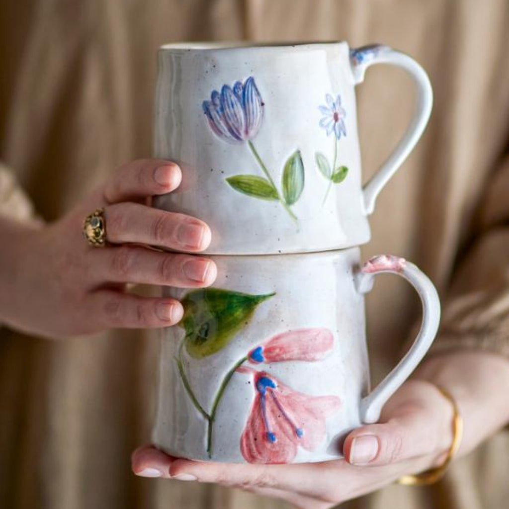 Two stacked ceramic mugs with floral designs held by a person against a beige background.