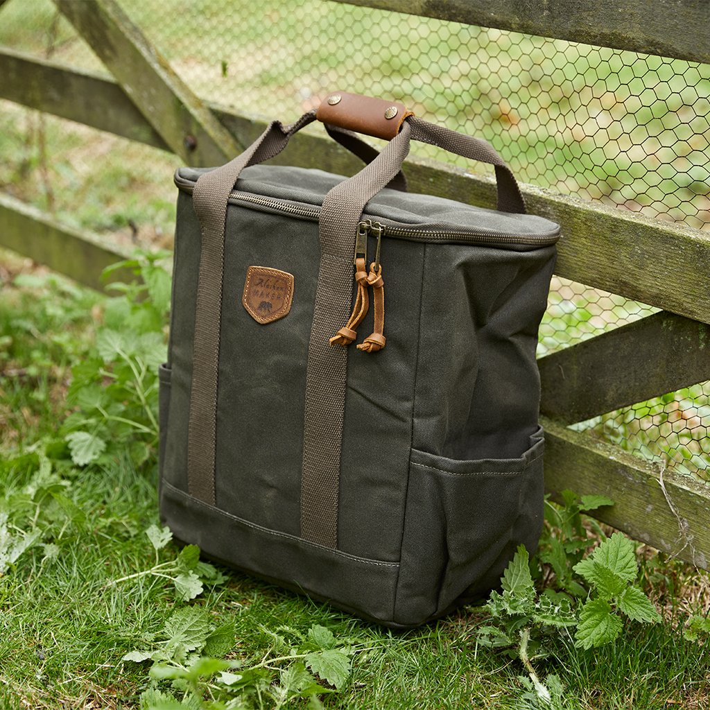 A dark olive green canvas cool bag with brown leather handles and zipper pulls, resting on grass in front of a wooden fence with wire mesh.