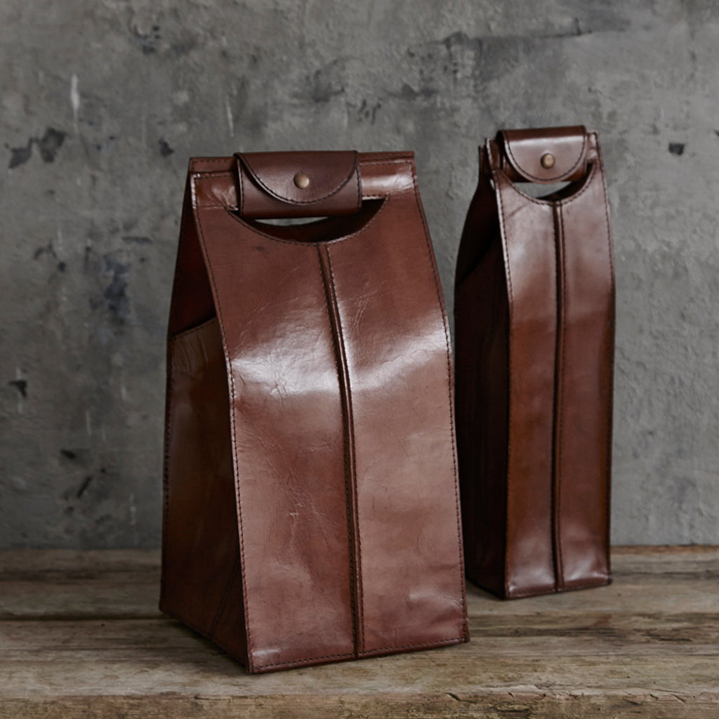 Two brown leather wine carriers with snap closures, displayed on a rustic wooden surface against a textured gray concrete wall.