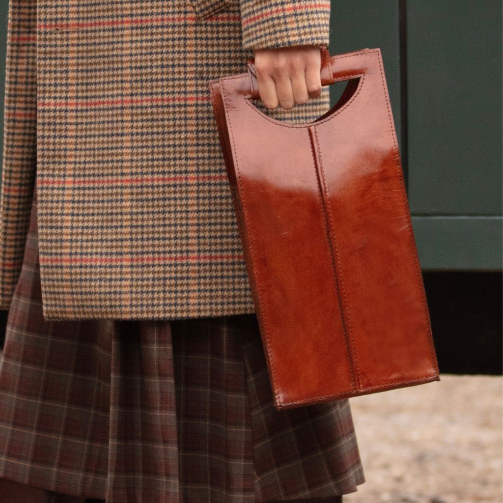 A person wearing a brown and beige plaid coat and skirt holding a leather wine carrier with a top handle.