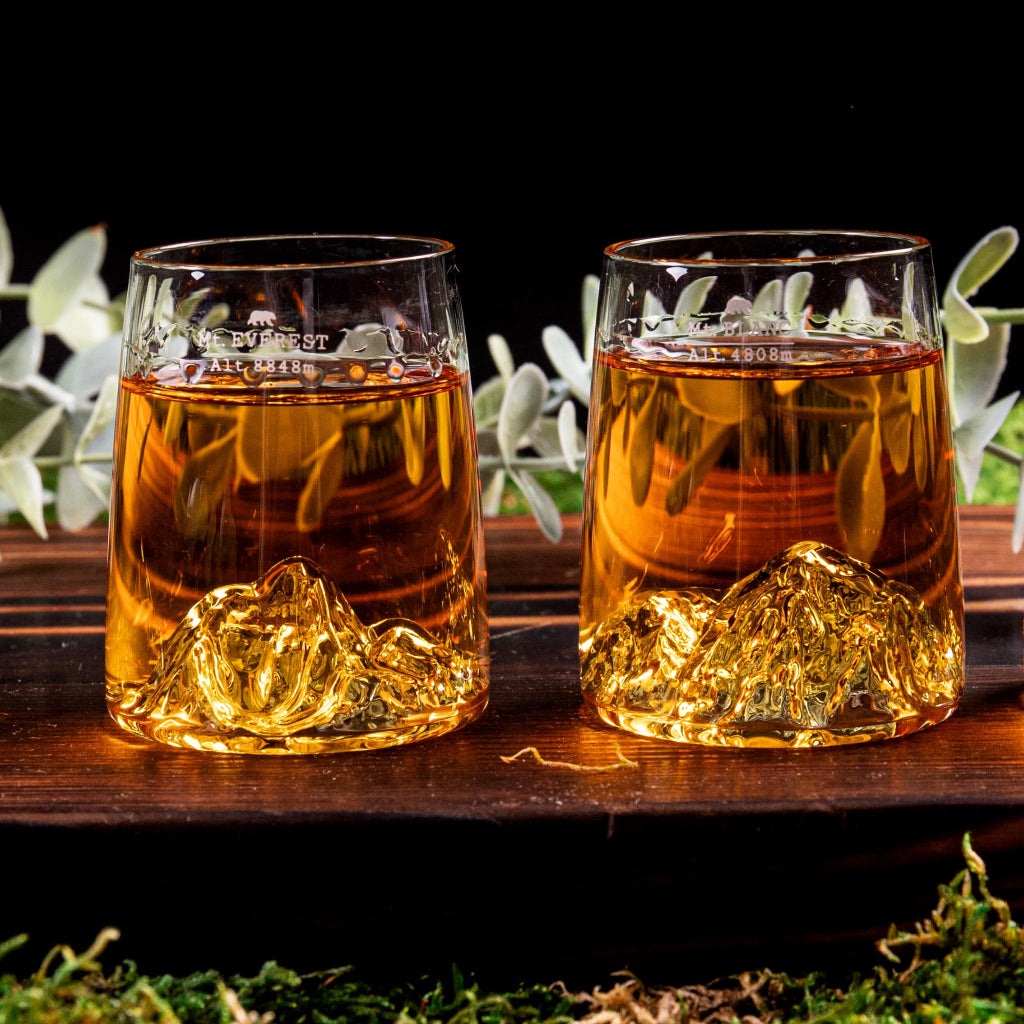 Two clear shot glasses with sculpted mountain-shaped bases, filled with amber liquid, displayed on a dark wooden surface against a black background with blurred greenery.