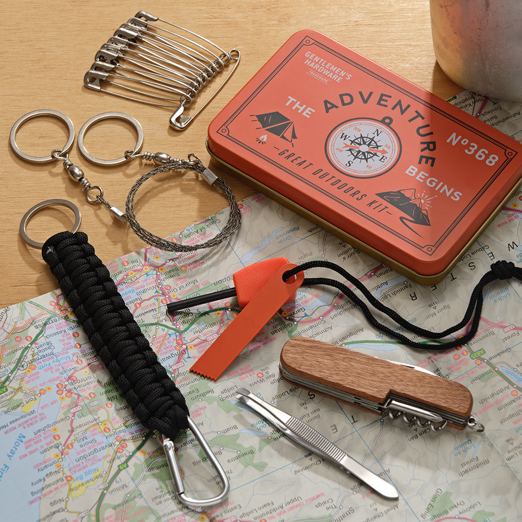 Collection of outdoor survival tools on a map with a 'Gentleman's Hardware' tin labeled 'The Adventure Begins'.