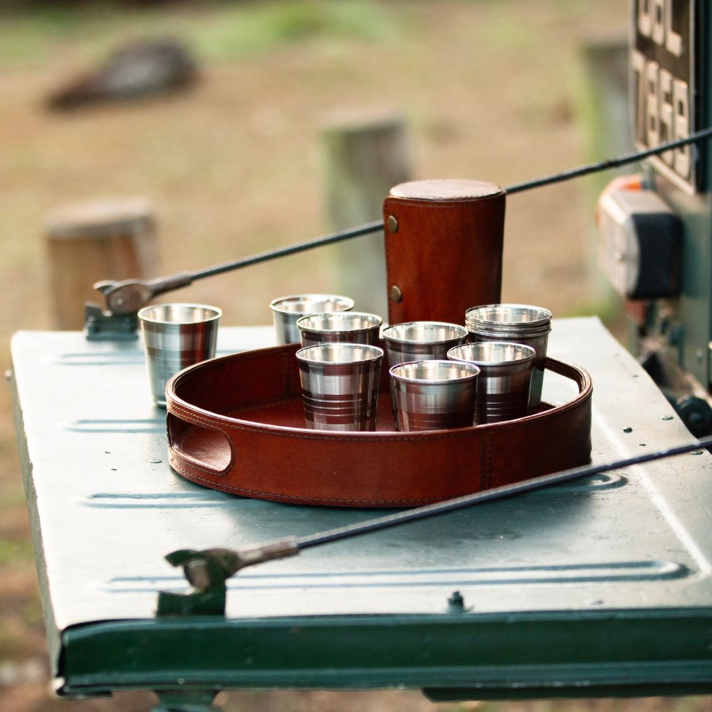 A set of six stainless steel shot glasses arranged in a brown leather tray, with a matching brown leather holder