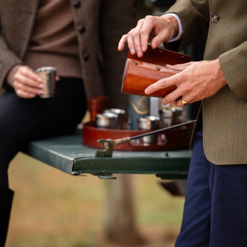 A close-up lifestyle shot of a person holding an open brown leather  holder for 10 stirrup cups, with another person seated beside them holding a metal cup. 
