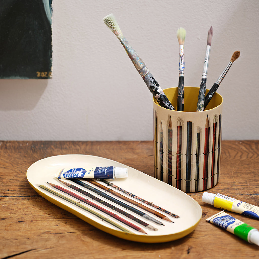 Art supplies including paintbrushes in a holder and tubes of paint on a wooden surface.