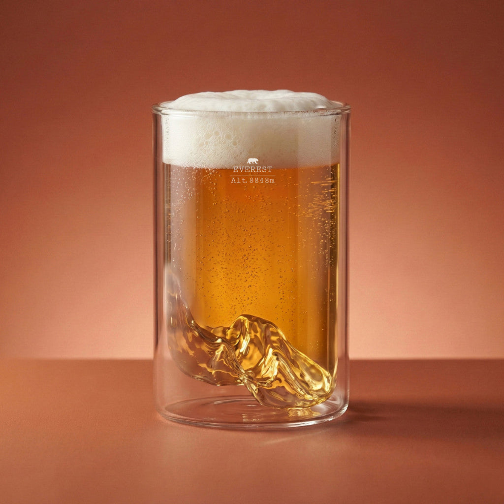 A clear double-walled glass mug filled with amber beer set against a warm terracotta background. The brand name 'EVEREST' is printed on the glass.