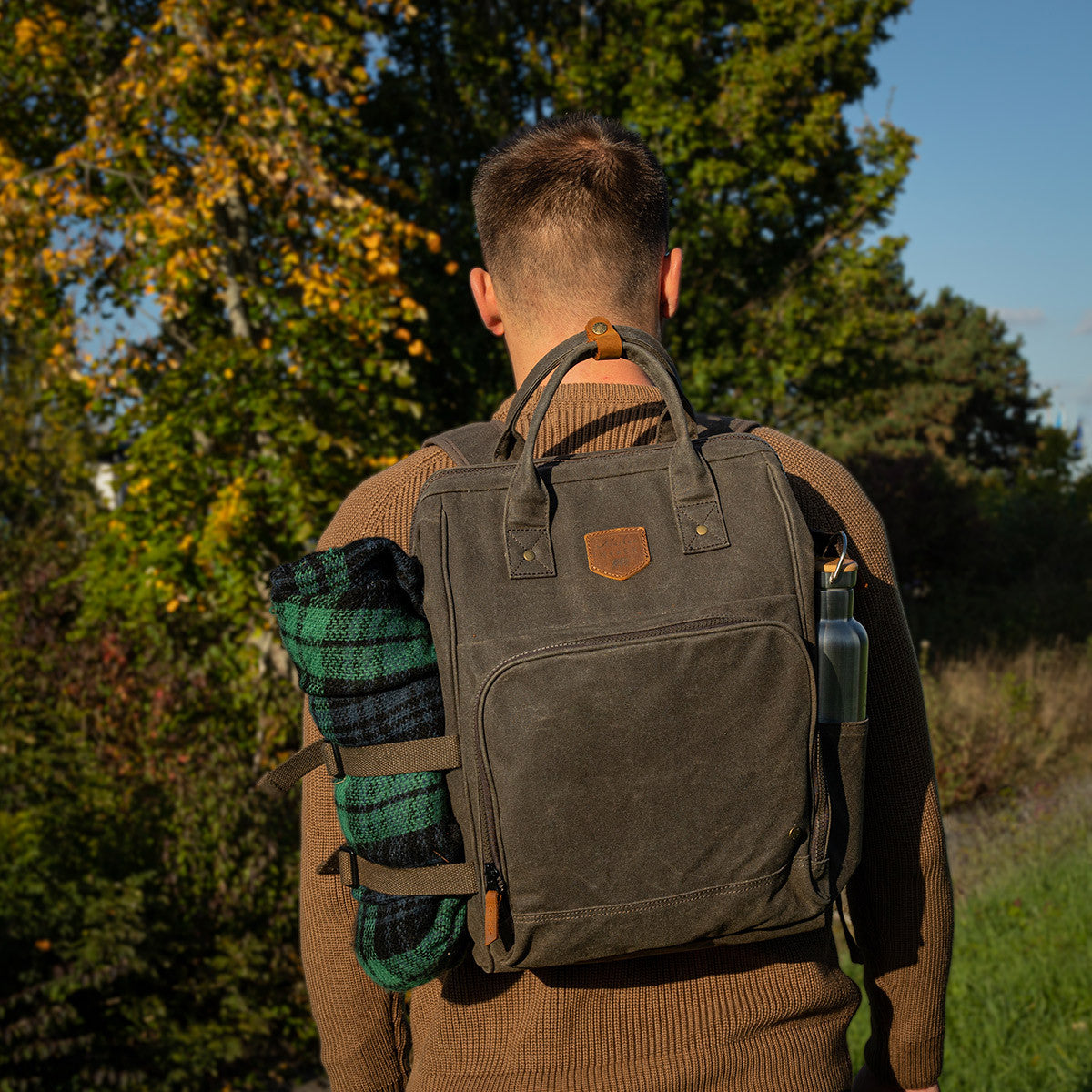 Waxed canvas picnic packpack cooler shown on the back of a man with picnic blacnket and water bottle.