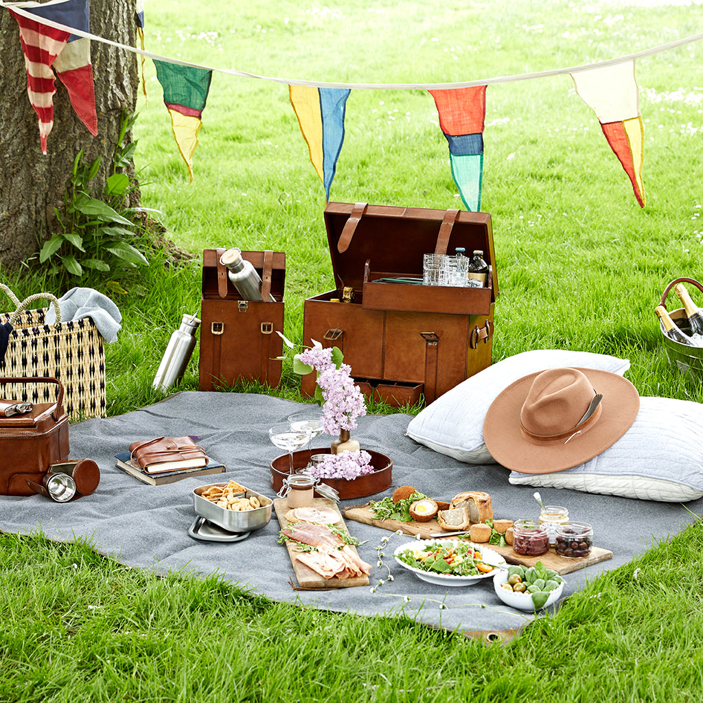 Waxed canvas and leather picnic rug with picnic set up including leather round drinks tray and picnic box with leather wine bottle carrier and leather cooler shown on picnic rug with cotton quilted grey and cream cushion covers