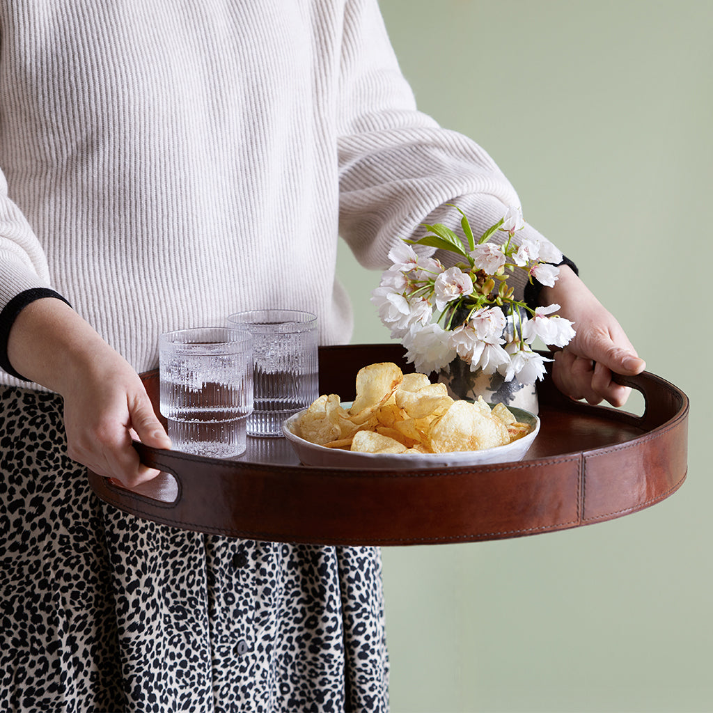 Leather serving tray with two cut o ut handles, the tray has a bowl of crisps, two glasses and a vase of flowers on it