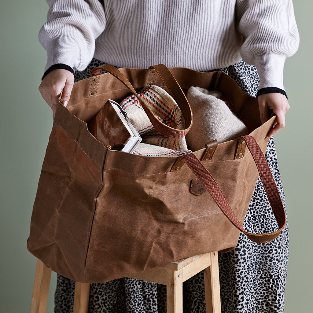 waxed canvas tote bag in brown, being held open with two blankets and a book